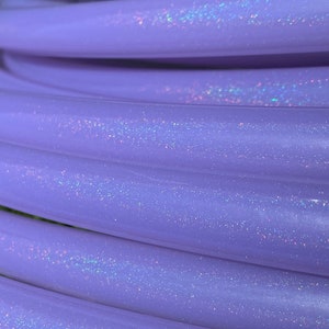 May include: Close-up of a coiled, lavender-colored hula hoop. The hoop has a smooth, slightly iridescent surface with scattered glitter particles. The image highlights the hoop's texture and color, suggesting a playful or fitness-related product.