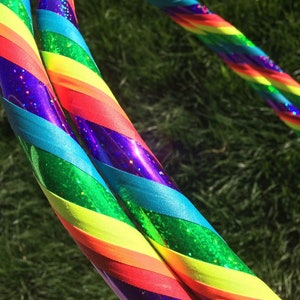 May include: Two rainbow-coloured hula hoops with glitter. The hoops are made of plastic and have a rainbow pattern with green, blue, yellow, orange, red, and purple stripes.