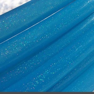 May include: Close-up of a vibrant blue material with a glossy finish. The fabric is folded, creating diagonal lines across the frame. Tiny, iridescent glitter particles are scattered throughout, catching the light and adding sparkle.