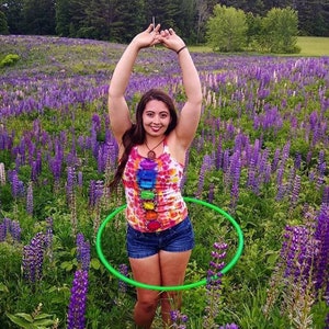 May include: A person wearing a tie-dye tank top with a rainbow pattern and denim shorts is holding a green hula hoop in a field of purple lupin flowers.