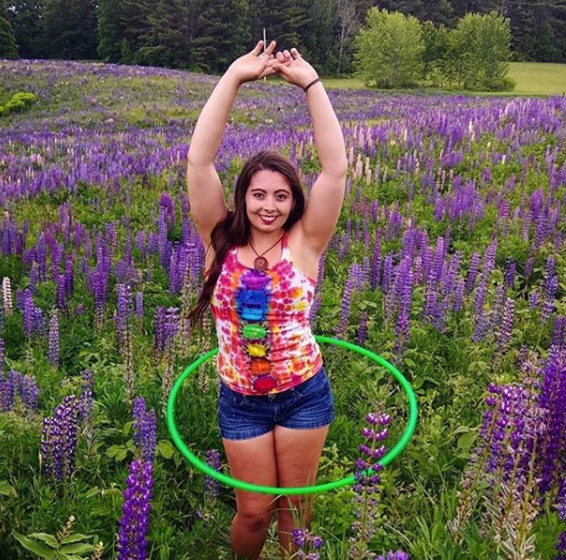 May include: A person wearing a tie-dye tank top with a rainbow pattern and denim shorts is holding a green hula hoop in a field of purple lupin flowers.