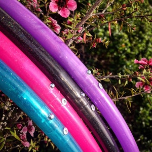May include: Four colorful hoop dance hoops with glitter and silver accents. The hoops are stacked on top of each other, with the purple hoop on top and the turquoise hoop on the bottom. The hoops are in front of a green leafy background with pink flowers.