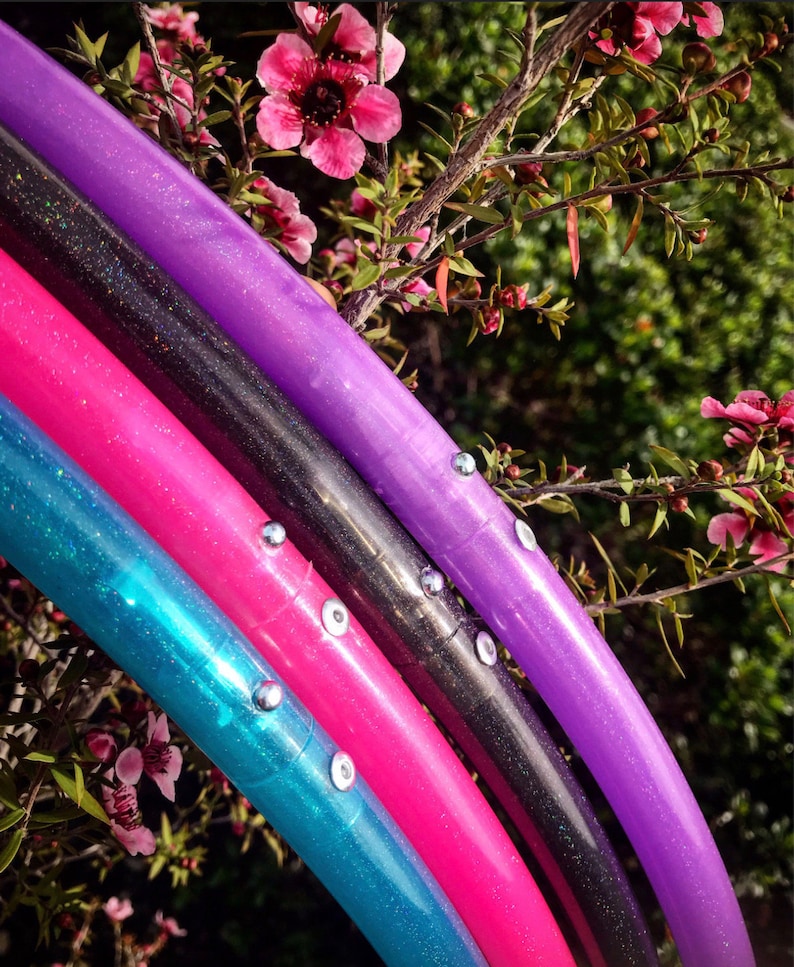 May include: Four colorful hoop dance hoops with glitter and silver accents. The hoops are stacked on top of each other, with the purple hoop on top and the turquoise hoop on the bottom. The hoops are in front of a green leafy background with pink flowers.