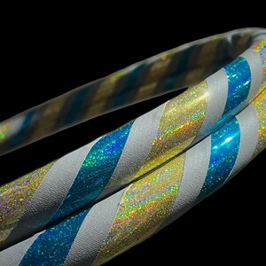 May include: Close-up of two iridescent hula hoops with a black background. The hoops have alternating stripes of silver, blue, and gold. The blue and gold stripes have a glittery, holographic effect.