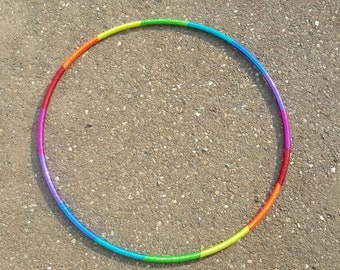 May include: A rainbow-colored plastic hula hoop on a gray gravel surface.