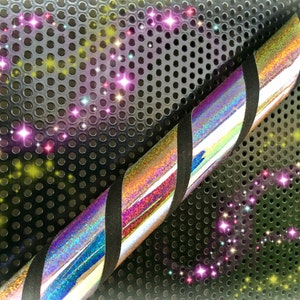 May include: A silver and black iridescent pole with a black grip. The pole is lying on a black metal surface with a pattern of holes. The background is blurred and has a sparkly effect.