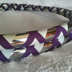May include: A silver hoop with purple and white chevron tape. The hoop is made of a flexible material and is designed for exercise or performance.