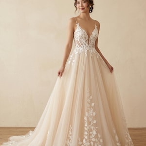 May include: A floor-length wedding dress featuring a beige tulle skirt and a bodice with a deep V-neckline. The bodice and the hem of the skirt are decorated with white floral lace appliqués. The dress has thin spaghetti straps and a full silhouette.