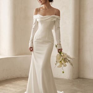 May include: A white off-the-shoulder wedding dress with long sleeves and a mermaid silhouette. The dress features a draped neckline and a fitted bodice. The bride is holding a bouquet of white flowers. The dress is floor-length.