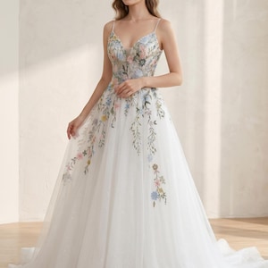 May include: A white wedding gown with a floral embroidered bodice and skirt. The dress features a V-neckline, spaghetti straps, and a flowing tulle skirt with delicate floral accents in pastel colors. The gown has a subtle sparkle.