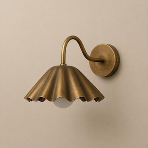 May include: A wall-mounted light fixture featuring a curved arm and a circular base plate in an antique brass finish. The lamp includes a wide, scalloped metal shade in the same brass tone, with a small, round, white glass bulb visible underneath.
