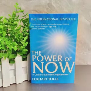 May include: A hardcover book titled "The Power of Now" with a blue cover and white text. The book is an international bestseller and a guide to spiritual enlightenment by Eckhart Tolle. A quote by Oprah Winfrey is also featured.