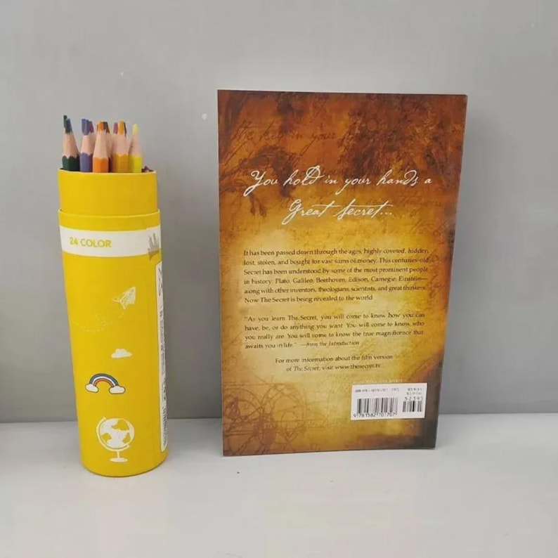May include: A yellow cylindrical pencil holder labeled "24 COLOR" next to a book with the title "The Secret." The pencil holder has a globe and rainbow design. The book's cover is brown with white text.