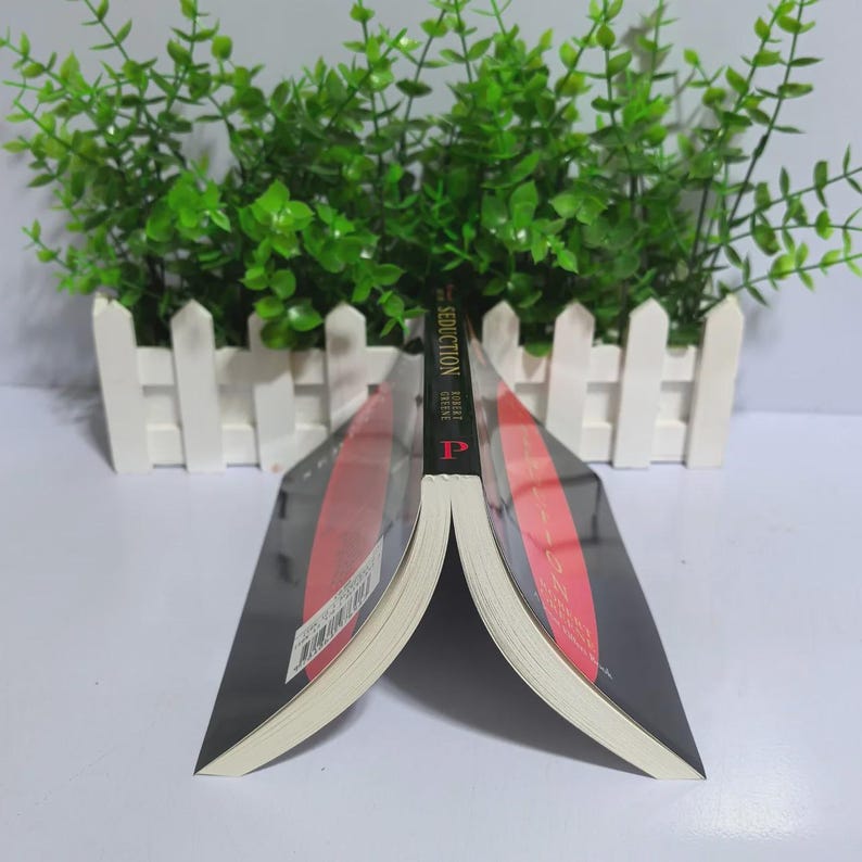 May include: An open book with a black cover and a red stripe design. The book's title, "Seduction", is visible on the spine. The book is positioned against a white picket fence and green foliage.