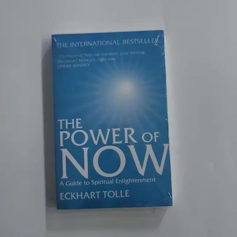May include: A blue book titled "THE POWER OF NOW" by Eckhart Tolle. The cover features the title in large white letters and a bright light emanating from the center. The book is an international bestseller and a guide to spiritual enlightenment.