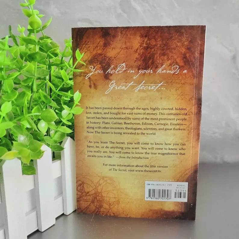 May include: A book cover with a textured, sepia-toned background. The title "The Secret" is displayed in elegant script. Text describes the book's themes of hidden knowledge and self-discovery, with references to historical figures.