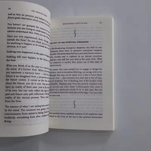 May include: Open book pages with text about the power of now. The text is in black font on a white background. The book is open to pages 50 and 51, with a decorative symbol on the right page.