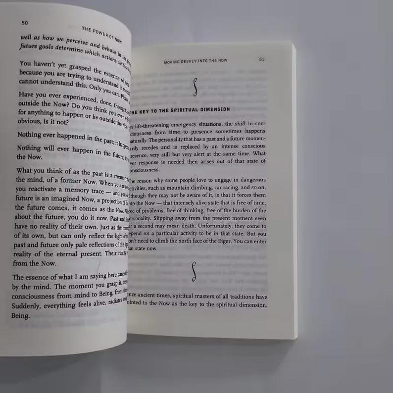 May include: Open book pages with text about the power of now. The text is in black font on a white background. The book is open to pages 50 and 51, with a decorative symbol on the right page.