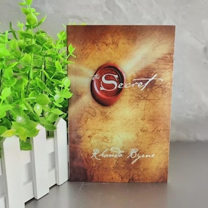 May include: A hardcover book titled "The Secret" by Rhonda Byrne. The cover features a red wax seal with the word "Secret" in white script, set against a parchment-like background with faded text.
