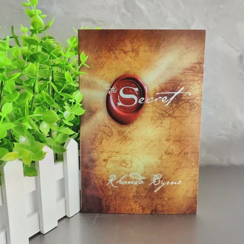 May include: A hardcover book titled "The Secret" by Rhonda Byrne. The cover features a red wax seal with the word "Secret" in white script, set against a parchment-like background with faded text.