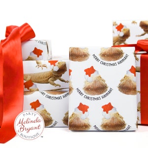 May include: A stack of white gift wrap with a red ribbon. The wrapping paper features a pattern of bearded dragons wearing Santa hats. The text "Merry Christmas Hannah!" is printed on the wrapping paper. The wrapping paper is from Melinda Bryant Boutique.
