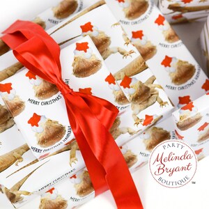 May include: White gift wrap with a repeating pattern of bearded dragons wearing Santa hats. The text "Merry Christmas Hannah" is printed on the wrapping paper. A red ribbon is tied around the gift boxes. The text "Party Melinda Bryant Boutique" is printed on a white circle.
