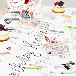 Table-length view of personalized butterfly coloring banner staged for a tea party. It is set with little cakes on miniature cake stands, shabby chic cups and saucers filled with crayons, and a teapot filled with red tulips.