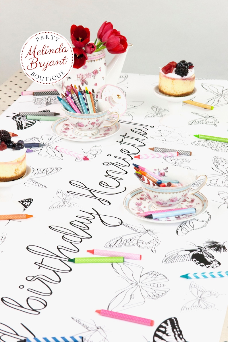 Table-length view of personalized butterfly coloring banner staged for a tea party. It is set with little cakes on miniature cake stands, shabby chic cups and saucers filled with crayons, and a teapot filled with red tulips.