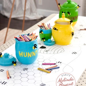 A table is set for a bee themed party or baby shower. A table length coloring page is laid over a dotted tablecloth. The design features happy bees, honeycombs, and a hive in the center. Optional text runs lengthwise along the edges. There are brightly painted honey jars filled with crayons on the table. This listing is for the coloring table runner only.