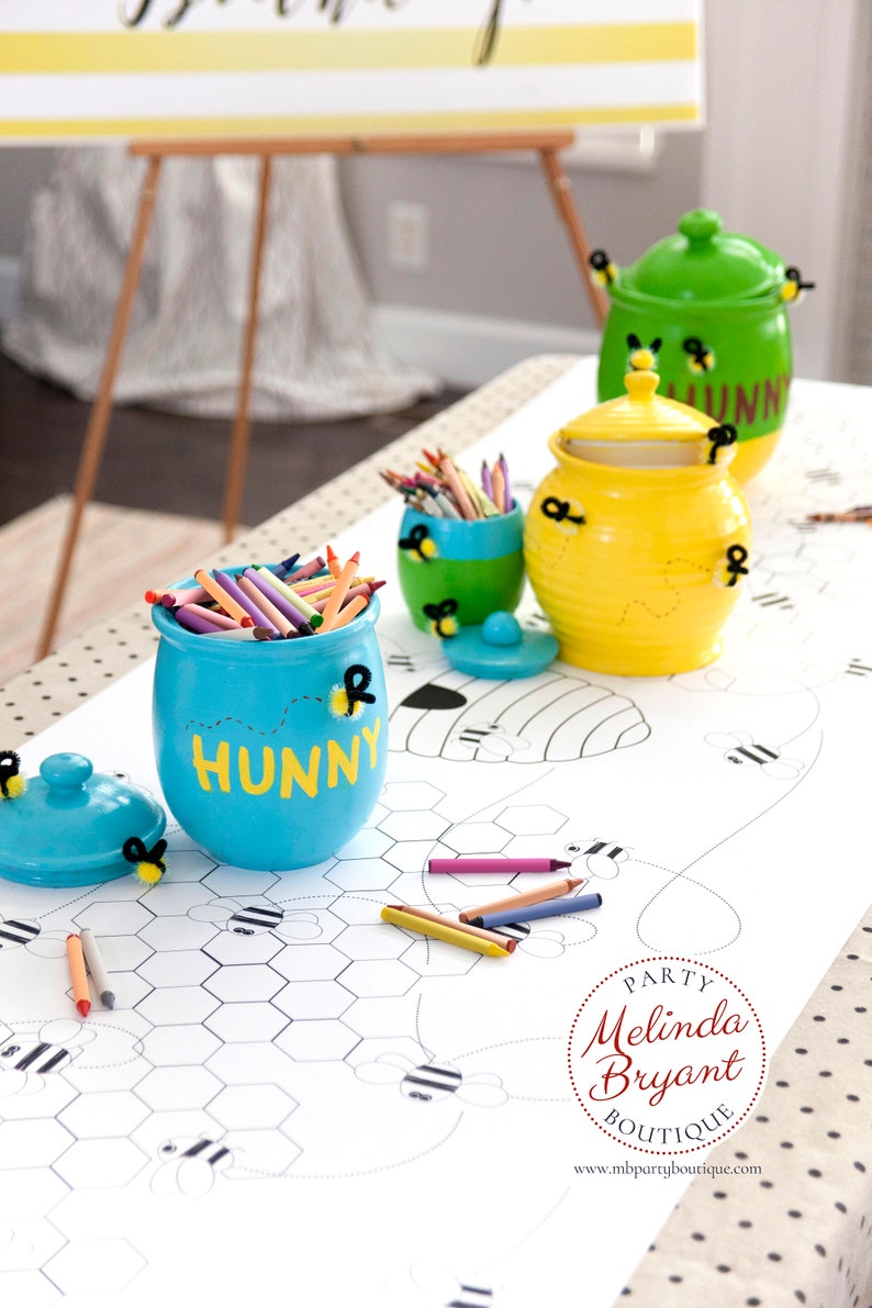 A table is set for a bee themed party or baby shower. A table length coloring page is laid over a dotted tablecloth. The design features happy bees, honeycombs, and a hive in the center. Optional text runs lengthwise along the edges. There are brightly painted honey jars filled with crayons on the table. This listing is for the coloring table runner only.