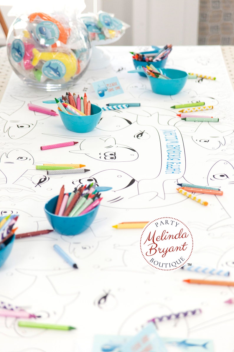 May include: A white table cloth with a shark-themed coloring page. The table is decorated with blue bowls filled with crayons and a glass bowl filled with small toys. The text "Happy 3rd Birthday Lily" is printed on the table cloth. The text "Party Melinda Bryant Boutique" is printed in a circle on the table cloth.