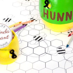 A table is set for a bee themed party or baby shower. A table length coloring page is laid over a dotted tablecloth. The design features happy bees, honeycombs, and a hive in the center. Optional text runs lengthwise along the edges. There are brightly painted honey jars filled with crayons on the table. This listing is for the coloring table runner only.