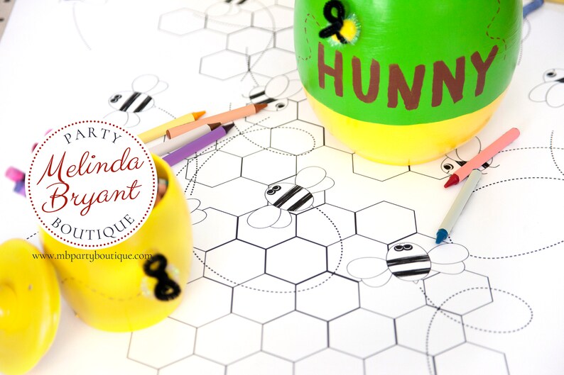 A table is set for a bee themed party or baby shower. A table length coloring page is laid over a dotted tablecloth. The design features happy bees, honeycombs, and a hive in the center. Optional text runs lengthwise along the edges. There are brightly painted honey jars filled with crayons on the table. This listing is for the coloring table runner only.