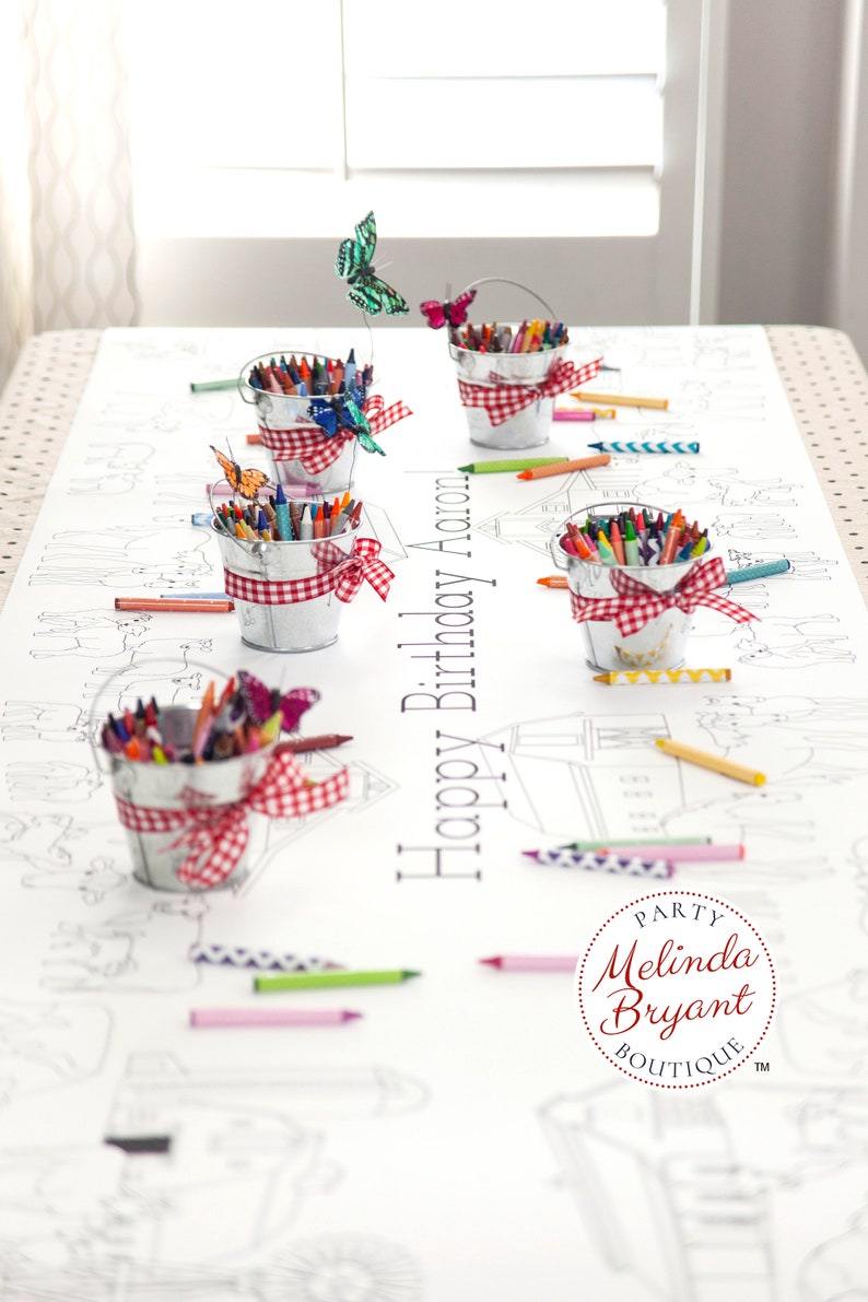 This table length view of my farm coloring table runner features horses, pigs, sheep, and a barn. The illustrated scene runs the length of the runner on both sides of the table. It is staged with miniature milk buckets filled with crayons.