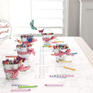 May include: A party table setting with small metal buckets filled with crayons, each adorned with a red and white gingham ribbon. The table runner features a coloring page design with the text "Happy Birthday". Butterflies decorate the scene.