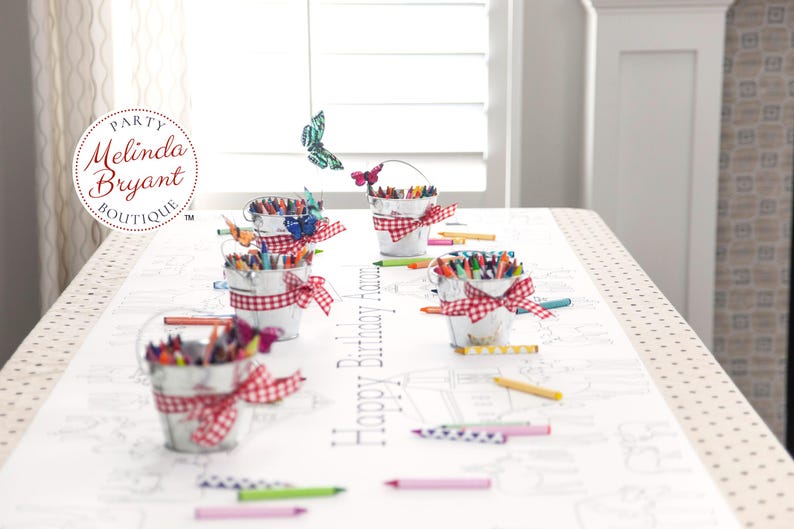 May include: A party table setting with small metal buckets filled with crayons, each adorned with a red and white gingham ribbon. The table runner features a coloring page design with the text "Happy Birthday". Butterflies decorate the scene.