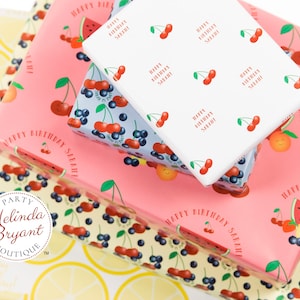 May include: Stack of three pink, white, and blue gift wrap with a lemon pattern on the bottom. The gift wrap features cherries, blueberries, watermelon, and the text "Happy Birthday Sarah".