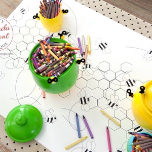 A table is set for a bee themed party or baby shower. A table length coloring page is laid over a dotted tablecloth. The design features happy bees, honeycombs, and a hive in the center. Optional text runs lengthwise along the edges. There are brightly painted honey jars filled with crayons on the table. This listing is for the coloring table runner only.