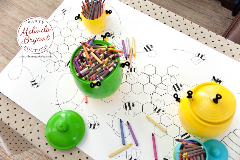 A table is set for a bee themed party or baby shower. A table length coloring page is laid over a dotted tablecloth. The design features happy bees, honeycombs, and a hive in the center. Optional text runs lengthwise along the edges. There are brightly painted honey jars filled with crayons on the table. This listing is for the coloring table runner only.