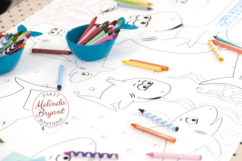 May include: A white table cloth with black outlines of sharks and other sea creatures. There are several bowls of crayons in various colors, including blue, green, red, and yellow. The table cloth has the text "PARTY Melinda Bryant BOUTIQUE TM" in a red circle.