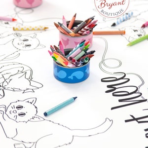 A white paper table cloth with black and white drawings of cats. There are three small repurposed tuna cans filled with crayons. The containers are decorated with a pink and white chicken design and a blue and white fish design (free printable labels available on my website).