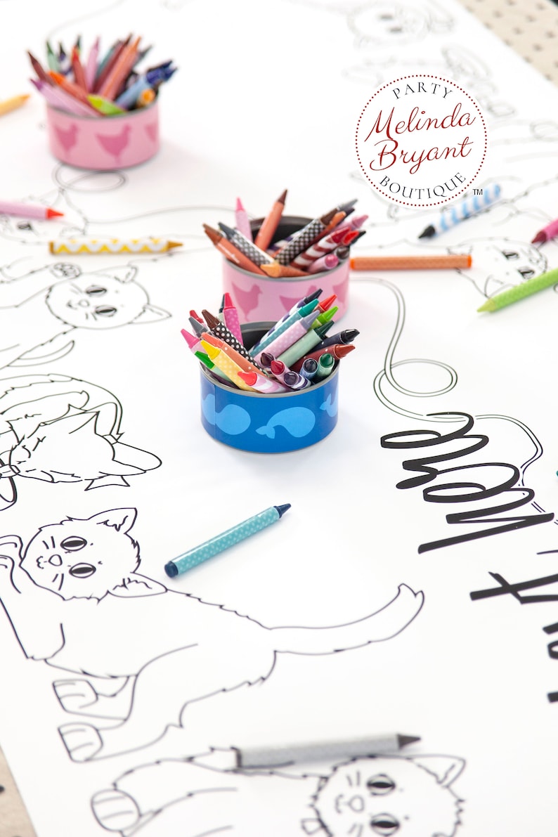 A white paper table cloth with black and white drawings of cats. There are three small repurposed tuna cans filled with crayons. The containers are decorated with a pink and white chicken design and a blue and white fish design (free printable labels available on my website).