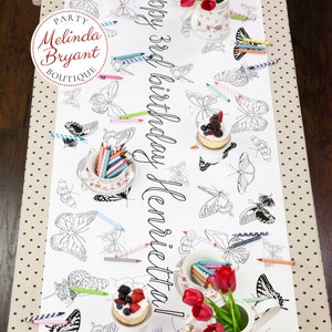 Table-length view of personalized butterfly coloring banner staged for a tea party. It is set with little cakes on miniature cake stands, shabby chic cups and saucers filled with crayons, and teapots filled with red tulips.