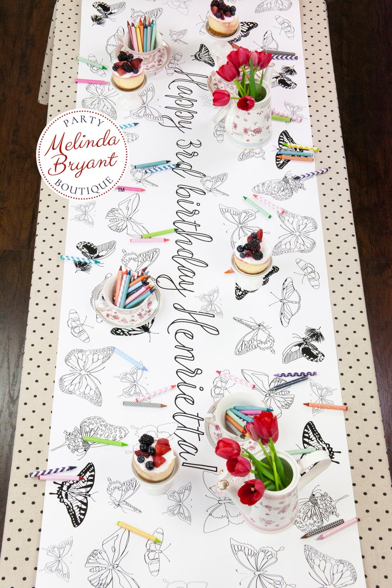 Table-length view of personalized butterfly coloring banner staged for a tea party. It is set with little cakes on miniature cake stands, shabby chic cups and saucers filled with crayons, and teapots filled with red tulips.