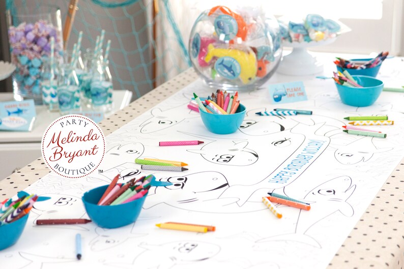 May include: A white table cloth with a shark-themed coloring page for kids. The table cloth has blue and white polka dots. There are several bowls of crayons in blue bowls on the table. The coloring page has the words "Happy Birthday" written on it.
