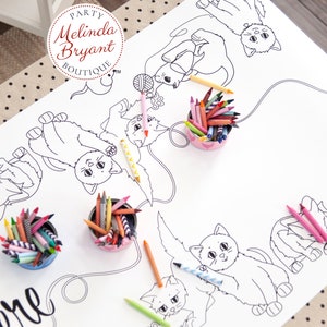 A table set with beige coloring table cloth with black polka dots. Over that, a paper table runner with black and white line drawings of cats playing with yarn and a mouse. There are four containers of crayons on the table cloth.