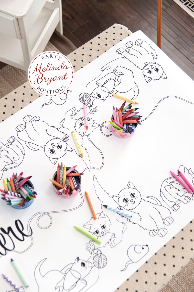 A table set with beige coloring table cloth with black polka dots. Over that, a paper table runner with black and white line drawings of cats playing with yarn and a mouse. There are four containers of crayons on the table cloth.