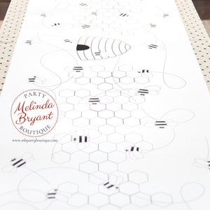 A table is set for a bee themed party or baby shower. A table length coloring page is laid over a dotted tablecloth. The design features happy bees, honeycombs, and a hive in the center. Optional text runs lengthwise along the edges. There are brightly painted honey jars filled with crayons on the table. This listing is for the coloring table runner only.