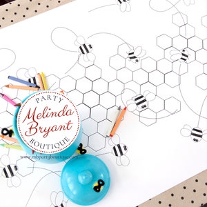 A table is set for a bee themed party or baby shower. A table length coloring page is laid over a dotted tablecloth. The design features happy bees, honeycombs, and a hive in the center. Optional text runs lengthwise along the edges. There are brightly painted honey jars filled with crayons on the table. This listing is for the coloring table runner only.