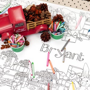 May include: A white coloring table cloth with a Christmas theme. The table cloth features a red toy truck with a pine cone and greenery in the bed. The table cloth also has a black and white illustration of presents, a rabbit, and a dog. The text "Bryant Family" is written on the table cloth. There are several crayons and colored pencils on the table cloth.